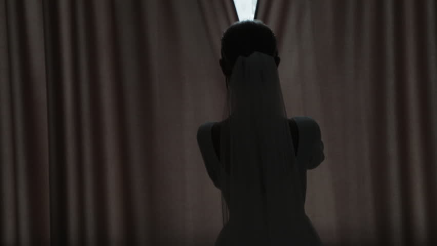 The silhouette of a beautiful bride in her wedding dress, standing in front of a window with closed curtains, sharply opens them, letting sunlight flood into the room.