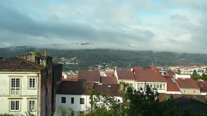 Rapid cloud movement crosses the rooftops of Santiago de Compostela as chimney smoke rises. Transition from shadow to sunlight while patches of fog and clouds move across the hills, changing weather.