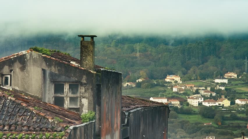 Fast-moving clouds sweep across the hills, as birds fly over an old rooftop and chimney in the foreground. Cars and distant people appear in the background while fog drifts across the trees