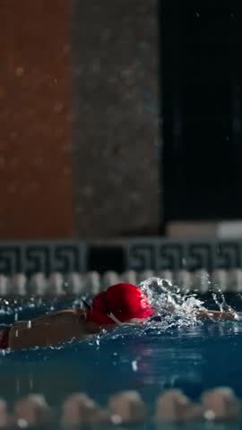 Focused swimmer in red cap powers through indoor pool lane, creating dramatic splashes and ripples in low light competitive training environment