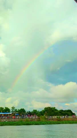 A vibrant rainbow arcs across a serene, cloudy sky above a tranquil body of water, with lush green trees and a distant shoreline creating a peaceful and picturesque natural landscape scene