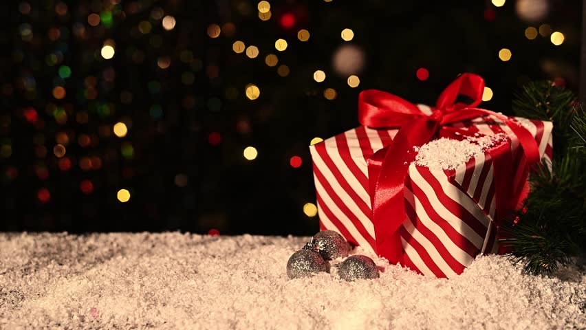 Christmas gift with red ribbon rests on snowy surface while background soft bokeh lights from Christmas tree create festive atmosphere and gentle illumination.Christmas background gift from copy space