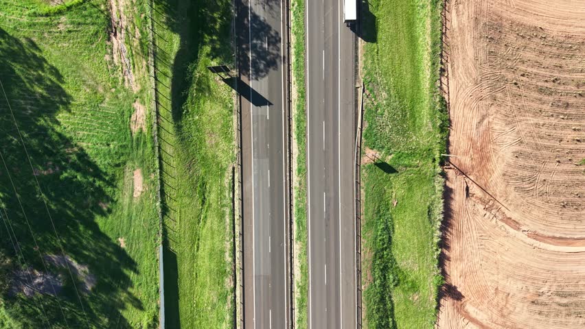 An aerial shot shows a truck driving on a highway. Lush green grass lines one side, while a freshly plowed field stretches out on the other. The sun casts shadows, adding depth to the scene.