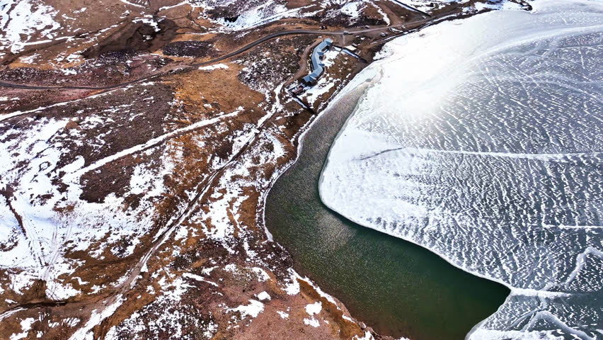 A snowy landscape with a river and a house in the distance. The sky is clear and the sun is shining brightly