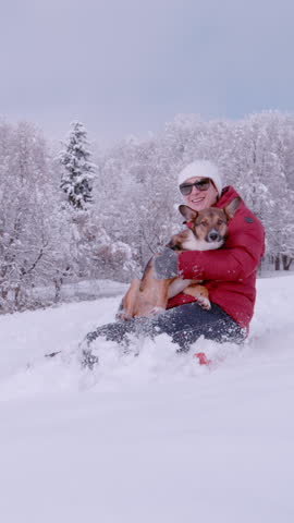 SLOW MOTION: Smiling lady is sledding on fresh snow with her cute dog in her arms. Cheerful woman having fun with her adorable furry friend while riding sled on a snowy hill on a beautiful winter day.