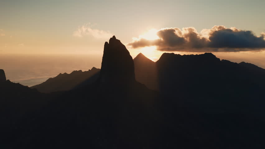 Moorea island mountains standing in silhouette against the warm glow of a tropical golden sunset, with sunbeams piercing through clouds over the Pacific Ocean