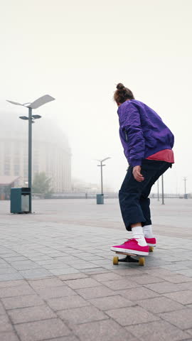 Rear view of young modern guy training his skateboarding skills in an empty city square on a foggy autumn morning outdoors. Find your freedom.