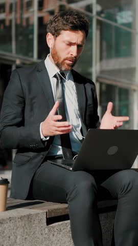 Businessman talks with partners on video conference via laptop sitting outdoors. Man in earphones explains specifics of project gesturing actively