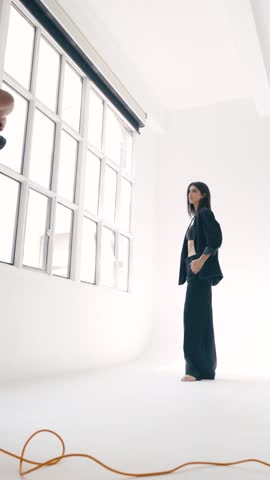Professional photographer capturing elegant female model posing in minimalist white studio setting during high-end fashion photoshoot. Vertical footage