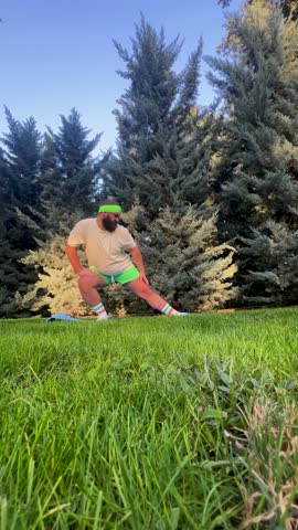 Street 4K loop video. Fat white man with beard, big belly, green shorts, t-shirt stands in surfer pose, stretching his legs and knees, preparing for exercise on blue mat on city park grass. Gym day