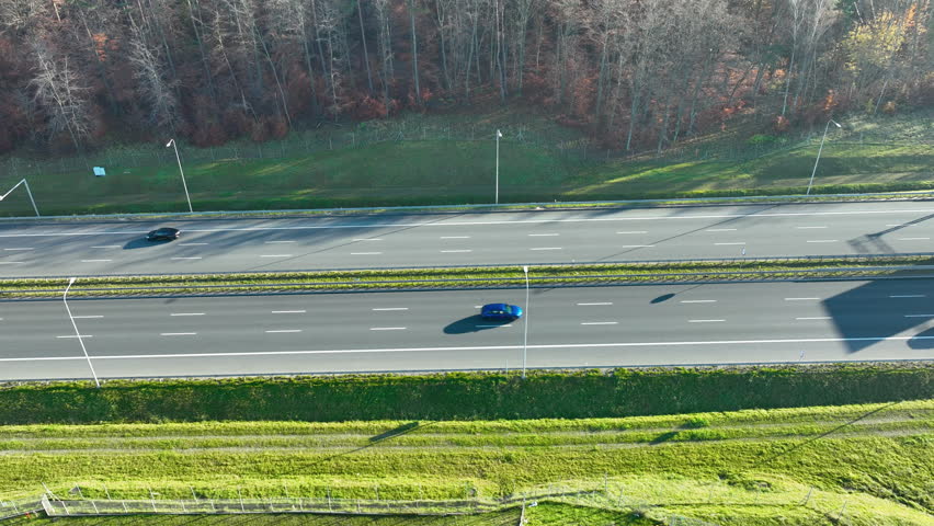Drone top-down view of a highway running beside a green verge and bare forest in daylight.