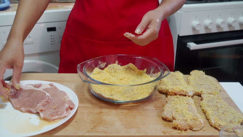 A cook in a red apron presses raw pork into yellow breadcrumbs in a glass bowl. She prepares the breaded meat on a wooden board in a home kitchen.