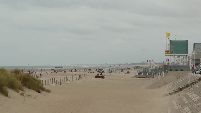Tractor moves along sandy beach promenade, cloudy sky, distant tourists, static wide shot, natural lighting