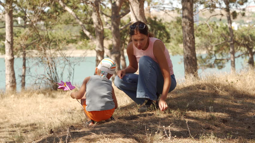 A woman and her son crouch on the dry grass to explore nature. The boy holds a pinwheel toy while they play in a sunny pine forest overlooking the lake.