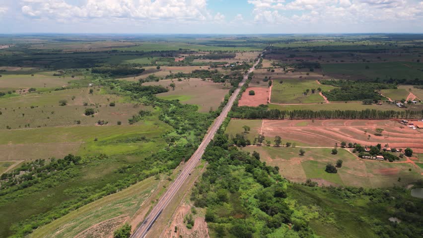 A wide aerial view of a long, straight highway or railway cutting through vast agricultural fields and natural riparian vegetation in the border region of São Paulo and Mato Grosso do Sul, Brazil.