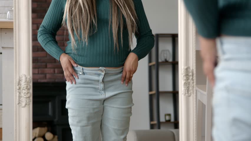 Close-up of overweight chubby black woman standing in front of mirror, struggling to fit into old tight pants after gaining weight while dressing up in domestic room.
