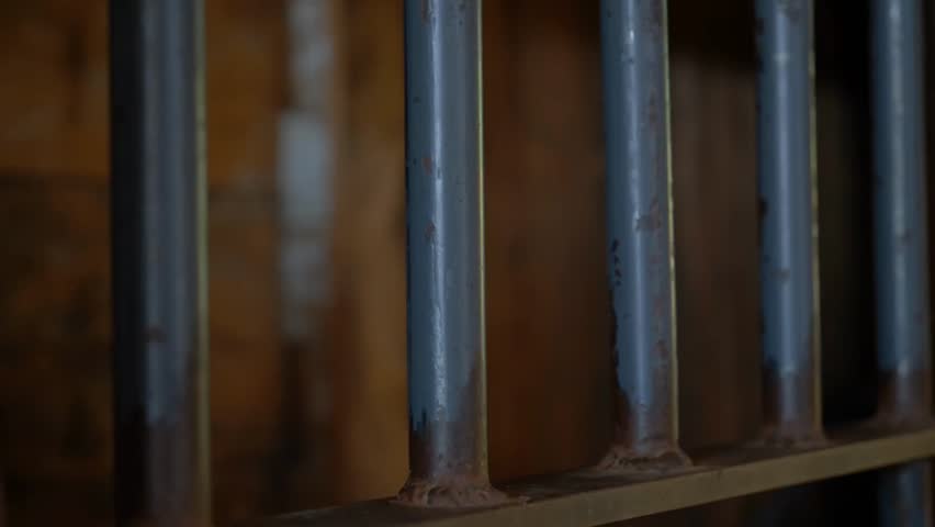 Close-up of a prisoner’s hands gripping metal bars inside a prison cell, depicting confinement, justice, and incarceration.