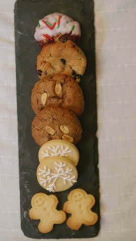 Variety of Christmas cookies on a rotating dark chalkboard, decorated with icing, sprinkles and festive figures in the foreground.