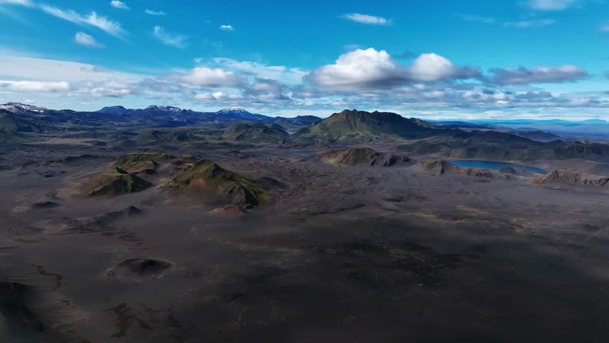 Aerial view over Iceland’s wilderness, revealing dark volcanic plains, scattered mossy hills, and distant lakes under bright blue skies and drifting clouds.