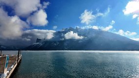 A snow dusted wooden bench faces a bright mountain lake, framed by bare and evergreen trees with sunlit water and hazy peaks in the distance, Alberta, Canada. - Powered by Shutterstock - Get 15% off with code: PIKWIZARD15