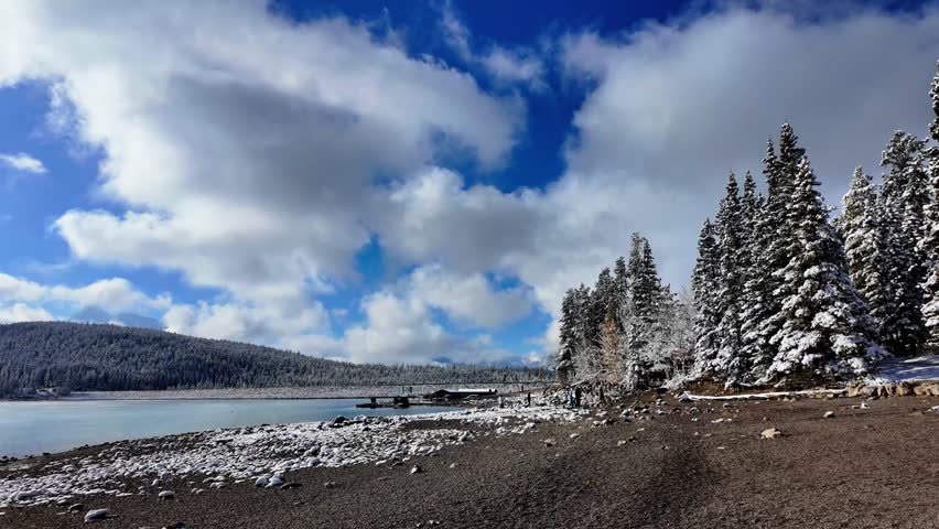 Wide view of a winter lake shoreline with snow covered rocks, distant wooded hills, and bright blue sky filled with soft clouds in the Canadian Rockies, Alberta, Canada.