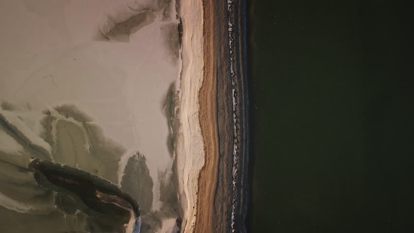 Camera glides forward above layered sand, snow, and ice forming clean lines and abstract patterns alongside dark, frozen water.