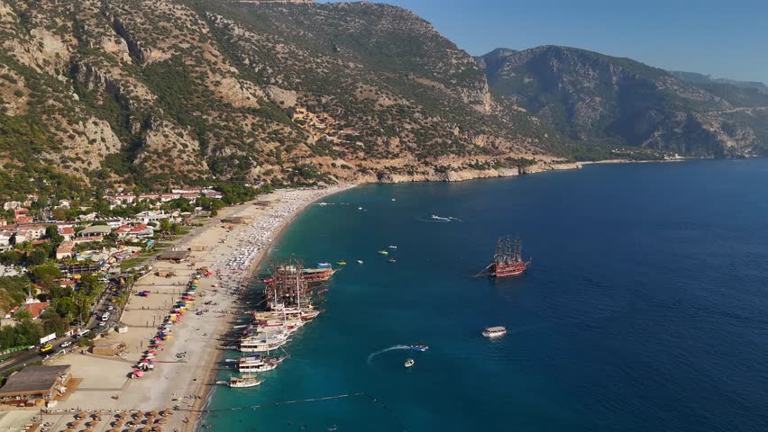 Aerial view of a long sandy beach with tour boats and pirate-themed ships near the coast in Turkey. Clear blue water, mountains, and summer activity create a vibrant coastal scene. Oludeniz, Turkey