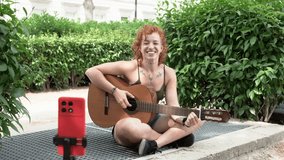 Cheerful female musician with red hair playing acoustic guitar and singing. Creative content creator vlogging or live streaming from a park - Powered by Shutterstock - Get 15% off with code: PIKWIZARD15