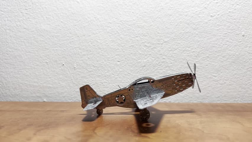 A small silver metal airplane model placed on a wooden surface with a white background. Close-up view highlighting the metallic texture and details of the miniature aircraft