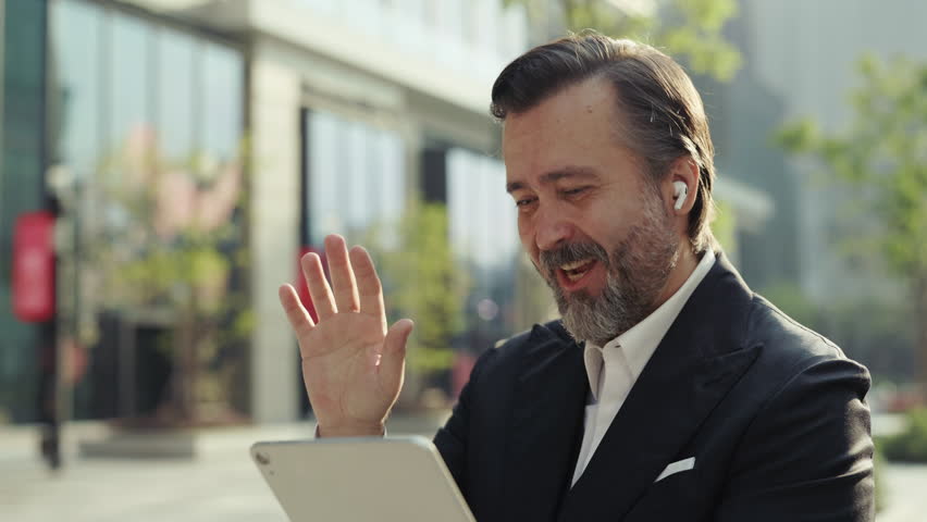 Portrait of elderly happy businessman waves hand Hello on video call via digital tablet outside office. Mature man entrepreneur joins online chat with coworkers on city street - Powered by Shutterstock - Get 15% off with code: PIKWIZARD15