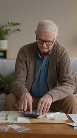 Vertical medium slowmo of distressed Caucasian senior man in glasses reviewing bills and counting expenses on sofa, managing personal finances and budgeting with concern and focus