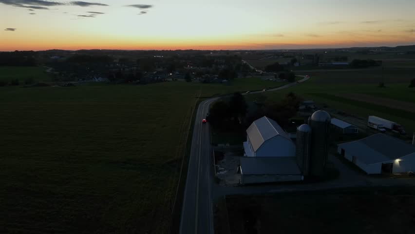 Headlights of cars on intersection during sunset time in USA. Aerial backwards wide shot. Silo storage of barn with house at dusk.