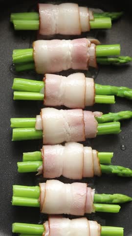 Raw bacon and asparagus rolls pan-fried with oil on a hot pan. Top view shot