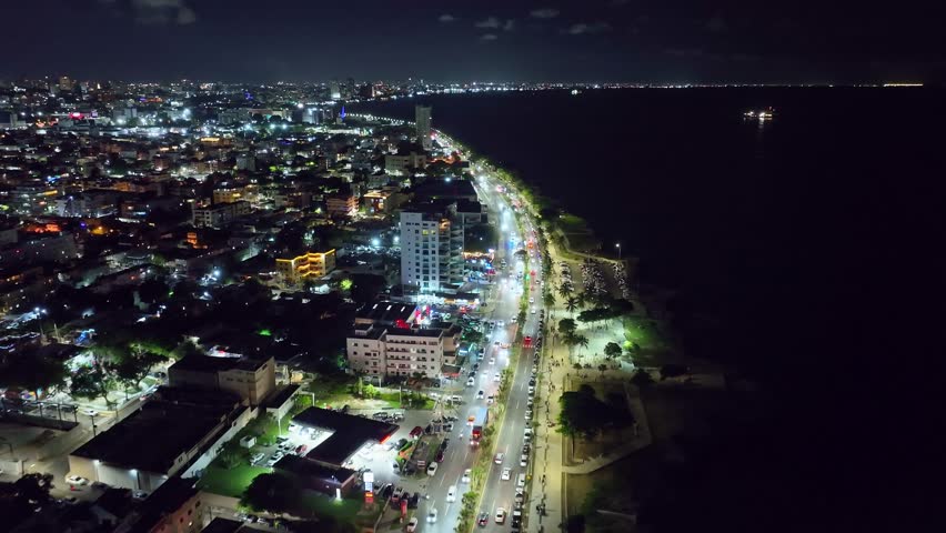 Cars on illuminated streets along coast of Santo Domingo city at night. Aerial Birds Eye shot. Illuminated city with apartment tower and houses in Dominican Republic. Wide shot. Peaceful scene.