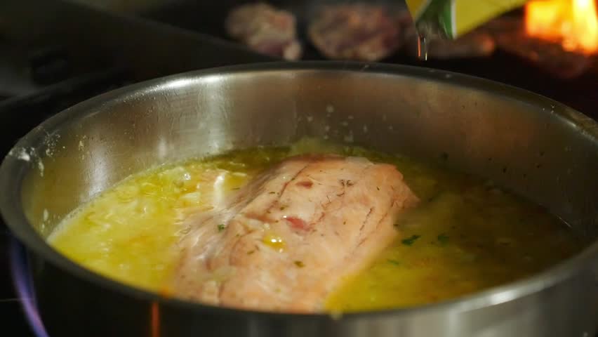 Chef pouring white wine over a salmon fillet in a pan with simmering sauce. Professional kitchen with grill and real fire in the background.
