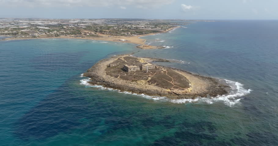 Aerial view of Isola delle Correnti, near Portopalo in Sicily, Italy. On the rocky island are the remains and ruins of an old building, once used as a barracks, and a lighthouse. Sunny summer day.