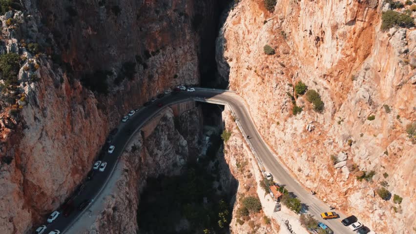 Aerial drone view showing winding canyon road and tunnel route cutting through rugged Saklikent Gorge in Mugla, capturing steep rock walls, moving traffic, narrow passage, and warm summer light