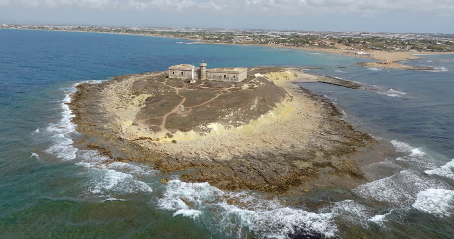 Aerial close-up of Isola delle Correnti, near Portopalo in Sicily, Italy. On the rocky island are the remains and ruins of an old building, once used as a barracks, and a lighthouse. Sunny summer day.