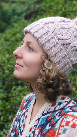 Young woman with curly hair wearing a knitted beanie and cozy cardigan feeling the cold weather outdoors, wrapping herself up while looking thoughtful and contemplative with a green background