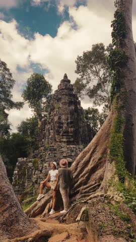 Young couple of travelers standing near giant tree roots in front of Bayon Temple face tower at Angkor Wat, Cambodia. Perfect for tourism, lifestyle promo, cultural heritage, and adventure footage.