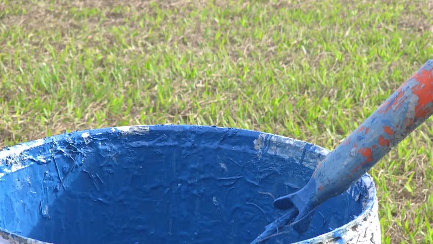 Vivid Blue Paint Dripping from Brush into Container on Grass Field in Bright Daylight