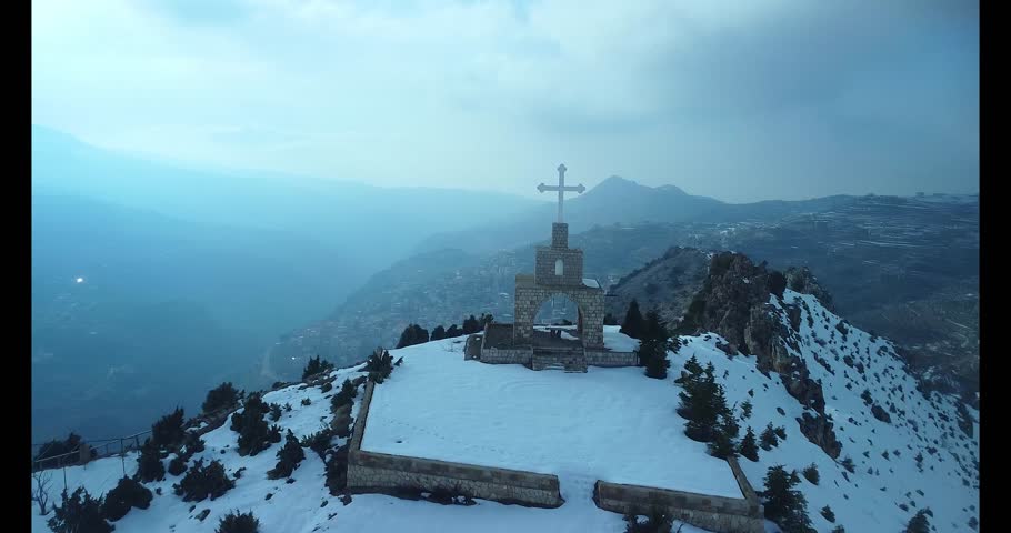 Aerial 4K cinematic orbit shot of a mountain cross overlooking the snowy Cedars of Bsharri in Northern Lebanon. The drone circles the historic landscape, revealing the iconic cedar forests, the cliffs