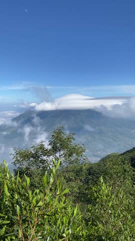 4k vertical video footage of Mount Sindoro in the distance from Mount Sumbing, Wonosobo, Indonesia with soft mist on the mountain slopes and blue sky.