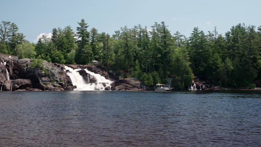 A pontoon boat near a powerful rocky waterfall surrounded by green forest and blue sky in High falls park, Bracebridge, Ontario, northern Canada