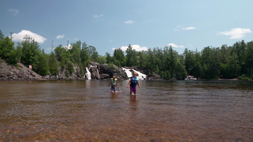 Two Caucasian children with life jackets on, walking in shallow water on a sand bank with a powerful waterfall in the background in High Falls Park