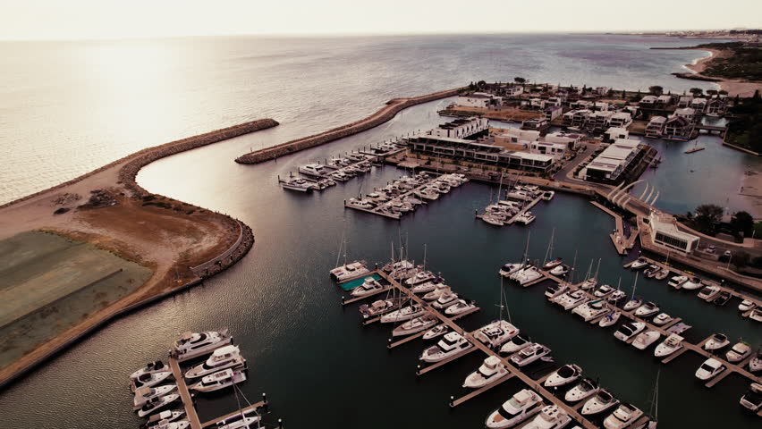 Aerial view of yachts are docked at the marina during sunset, with calm waters and a coastal landscape in the background.