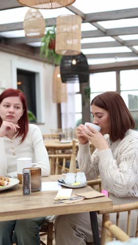 Two friends sit in a cafe and one sips her coffee then instantly grimaces at the terrible taste struggling to swallow while the other watches surprised trying to understand what went wrong