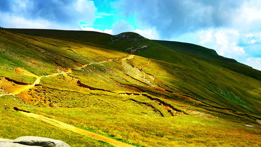 Winding dirt path across green and yellow slopes under partly cloudy sky. Peaceful hiking trail destination. Romanian mountain.