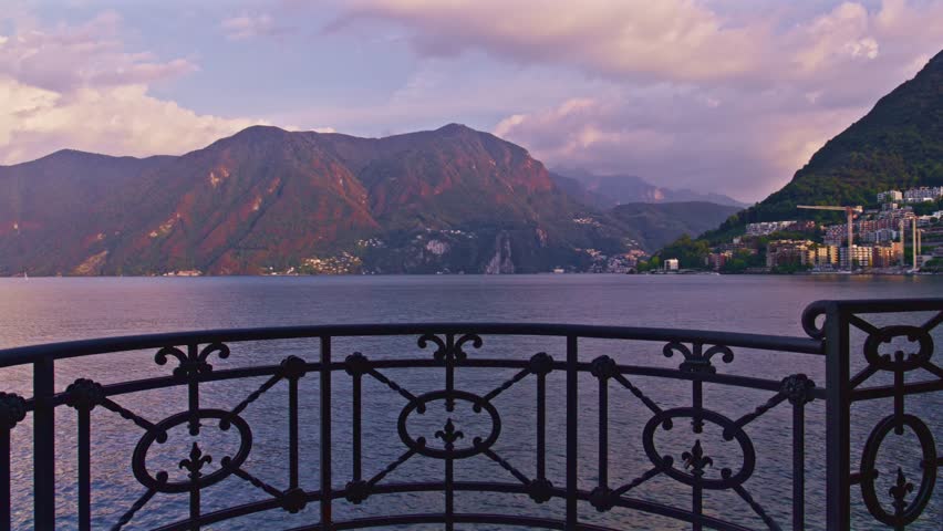 Lugano, Switzerland – October 5, 2022: View across Lake Lugano toward city of Paradiso, Monte San Salvatore and Monte Caprino, framed by decorative metal railing in the foreground.