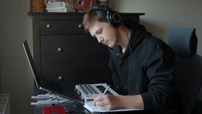 Focused songwriter wearing studio headphones pauses digital arrangement on laptop to write notes and compositional ideas in paper notebook, blending analog planning with technology in home studio - Powered by Shutterstock - Get 15% off with code: PIKWIZARD15
